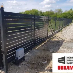 Automatyczna brama przesuwna z poziomymi panelami, widoczny napęd i fragment ogrodzenia z siatki, teren przed posesją w trakcie prac ziemnych.