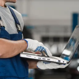 Mechanik w niebieskim kombinezonie i roboczych rękawicach, trzyma otwarty laptop w warsztacie samochodowym; w tle widoczny podnośnik i fragment naprawianego pojazdu.