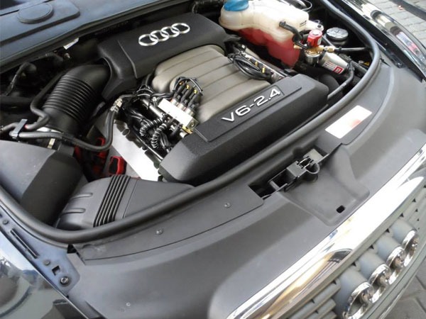 Otwarta maska samochodu Audi, widoczny silnik V6 2.4 z elementami instalacji gazowej, zbiornik płynu chłodniczego i przewody.