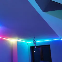 Wnętrze pokoju z zamontowanym oświetleniem LED RGB na suficie i ścianach, tworzącym wielokolorowe efekty świetlne, widoczna surowa żarówka wisząca na kablu.