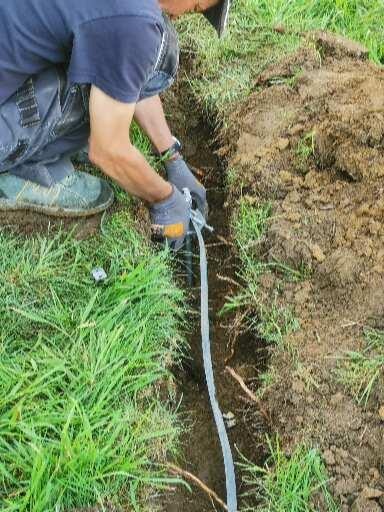 Elektryk w trakcie układania kabla w wykopie ziemnym, zabezpieczonego szarą rurą karbowaną, widoczne narzędzia i fragment trawnika.