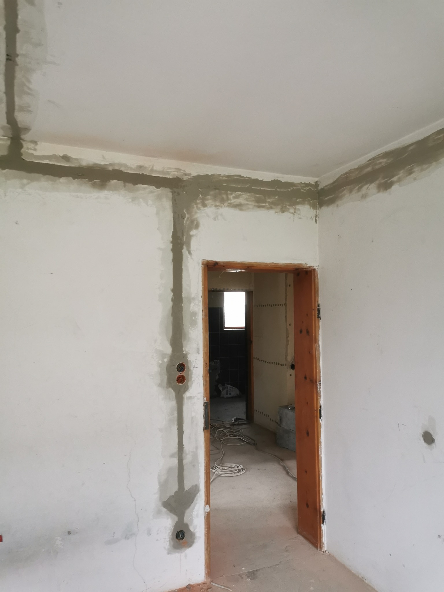 Wnętrze w trakcie prac wykończeniowych: ściany z bruzdami pod kable elektryczne, z widocznymi puszkami instalacyjnymi, częściowo zamaskowanymi gipsem, surowa ościeżnica drzwiowa.