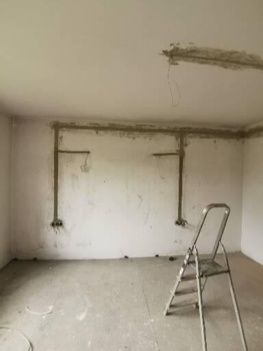 Wykute bruzdy pod instalację elektryczną w surowym pomieszczeniu, widoczne przewody i puszki, drabina oparta o ścianę, ślady gipsu na suficie.