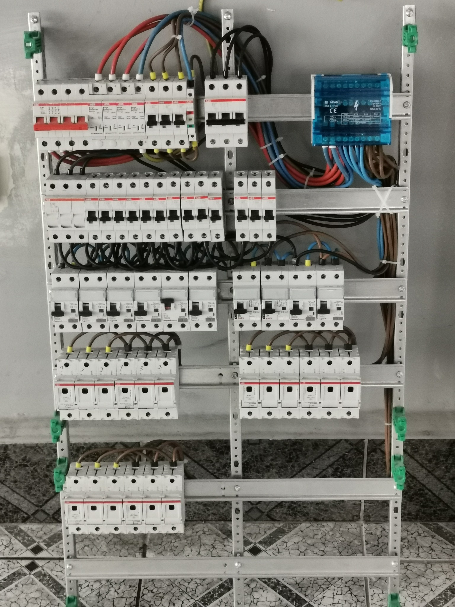 Rozdzielnica elektryczna z licznymi zabezpieczeniami nadprądowymi i różnicowoprądowymi, widoczne przewody w różnych kolorach podłączone do poszczególnych modułów, całość zamontowana na metalowej...