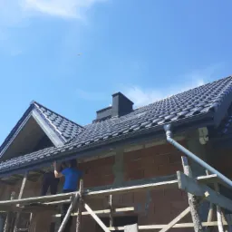 Budowa domu murowanego: murarze na rusztowaniu montują rynny pod nowym dachem z dachówki ceramicznej w słoneczny dzień.