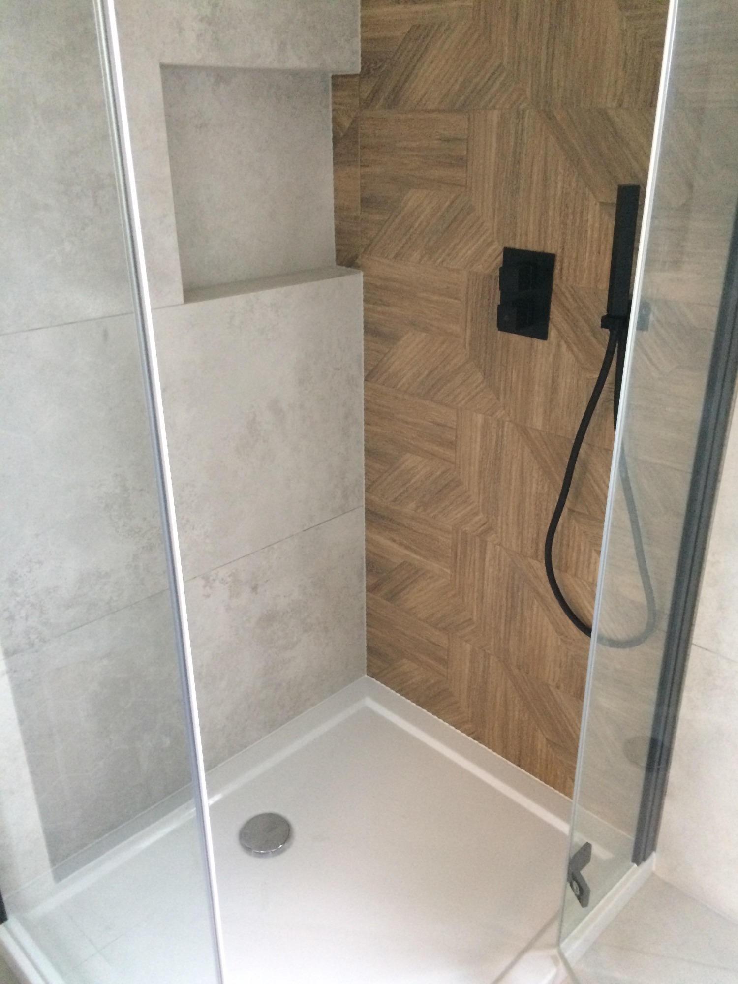 Kabina prysznicowa typu walk-in z odpływem liniowym i półką w ścianie, wykończona płytkami w odcieniach szarości i drewnopodobnymi z brązowym wzorem jodełki na ścianie z czarną armaturą.