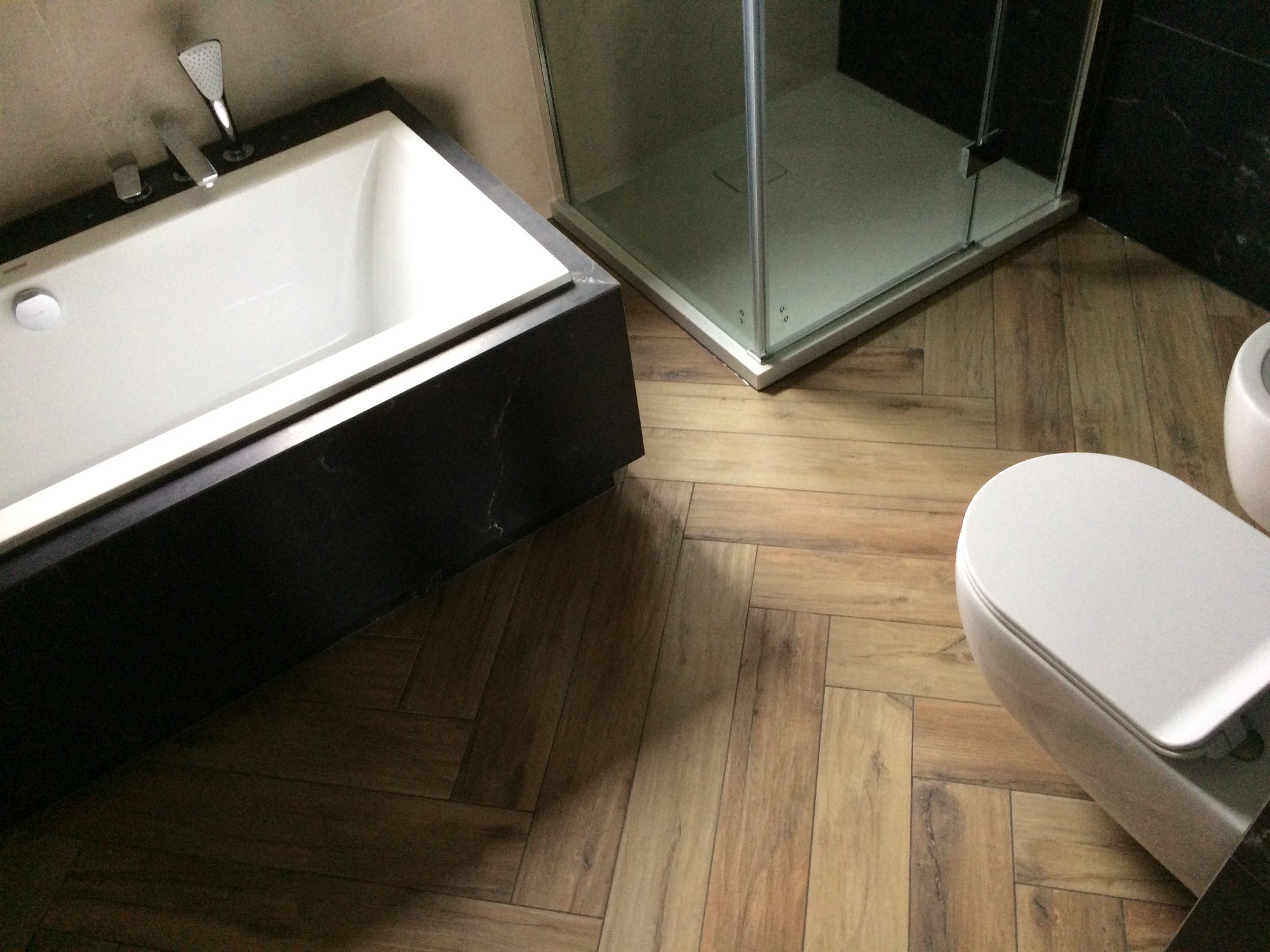 Nowoczesna łazienka z białą wanną obudowaną czarnym kamieniem, kabiną prysznicową ze szkła i toaletą, wykończona drewnopodobnymi płytkami ułożonymi w jodełkę.