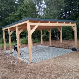 Drewniana wiata w trakcie budowy, z widocznymi słupami, konstrukcją dachu i częściowo ułożoną kostką ażurową na podłożu, otoczona ziemią i zielenią.