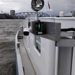 Biała kamera monitoringu oraz lampa nawigacyjna ze świecącym na zielono światłem, zamontowane na burcie statku z widokiem na rzekę i most w tle.