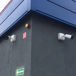 Dwie kamery monitoringu, sygnalizator alarmowy i zielona strzałka kierunkowa zamontowane na szarej, teksturowanej ścianie budynku z niebieskim dachem.