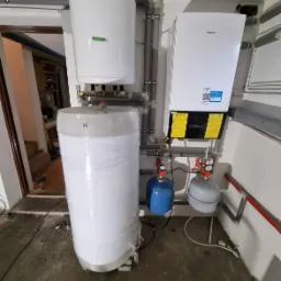 Biały zbiornik buforowy owinięty folią, podłączony do kotła gazowego Daikin, z niebieskim i szarym naczyniem przeponowym, widoczne rury i złączki, surowy beton na podłodze, widok na pomieszczenie...