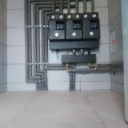 Szare rury instalacyjne prowadzone wzdłuż ściany wyłożonej białymi płytkami, z trzema czarnymi skrzynkami kontrolnymi i mosiężnymi elementami złącznymi. Widok z dołu, ukazujący fragment podłogi...