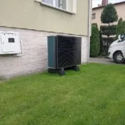 Zewnętrzna jednostka pompy ciepła ustawiona na trawniku obok budynku z jasną elewacją i białą skrzynką elektryczną. W tle widoczny biały samochód dostawczy z otwartymi drzwiami.