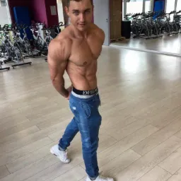 Mężczyzna o wysportowanej sylwetce, w dżinsach i sportowych butach, pozuje w siłowni z rowerami spinningowymi w tle, z widocznym logo marki na pasku.