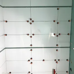 Ściana wyłożona białymi, prostokątnymi płytkami z użyciem systemu poziomowania płytek z pomarańczowymi klipsami, widoczne fugi i narzędzia glazurnicze w dolnej części kadru.