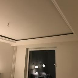 Lux-Bud Łukasz Wójtowicz - Fragment pokoju z białym sufitem z dekoracyjnymi listwami, widoczne okno z odbiciem lamp i łóżka, oraz wystające kable ze stropu.