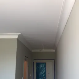 Pomalowane na szaro ściany z białym, ozdobnym wykończeniem sufitu, zabezpieczone folią drzwi w tle.