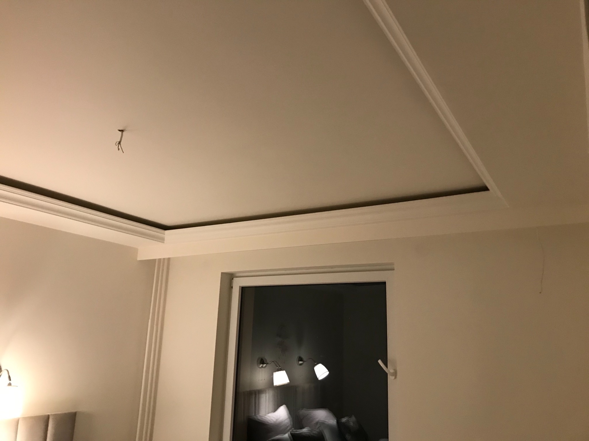 Fragment pokoju z białym sufitem z dekoracyjnymi listwami, widoczne okno z odbiciem lamp i łóżka, oraz wystające kable ze stropu.