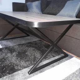 Stolik kawowy z blatem imitującym drewno i metalową podstawą w kształcie litery X, ustawiony na wzorzystym dywanie obok sofy z poduszkami.