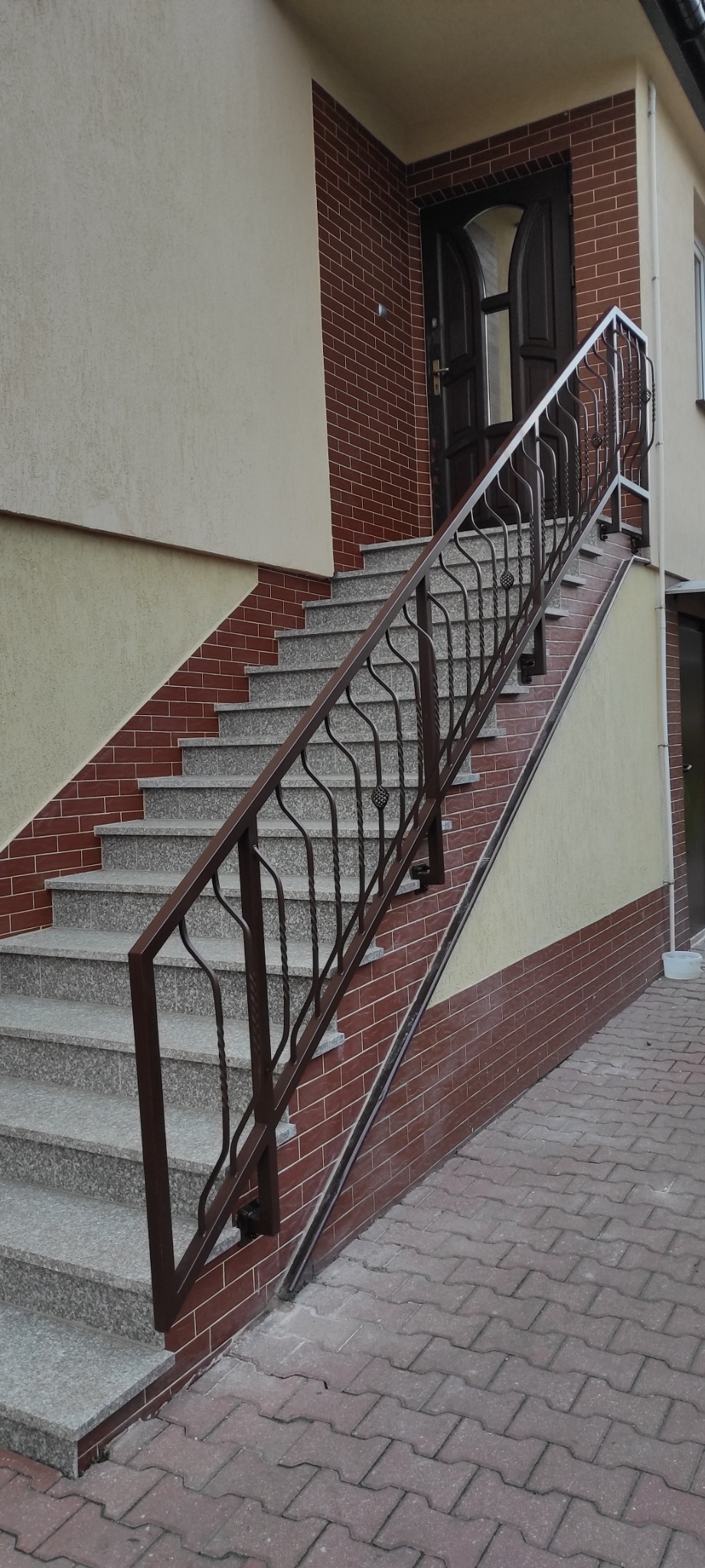 Zewnętrzne schody z szarego kamienia z brązową, metalową poręczą o ozdobnym wzorze, prowadzące do drzwi wejściowych w budynku z ceglaną elewacją.