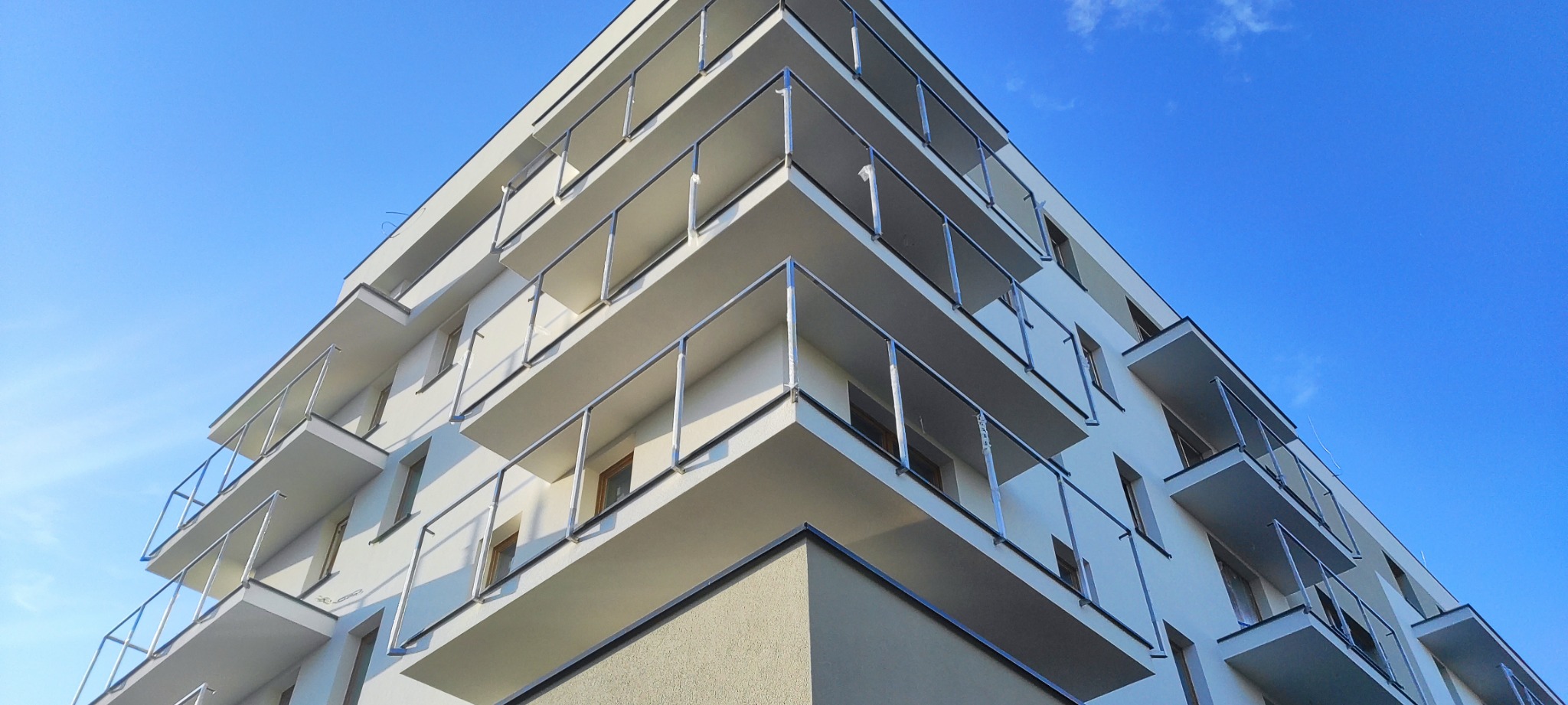 Nowoczesny budynek mieszkalny z balkonami zabezpieczonymi szklanymi balustradami w stalowych ramach, widziany pod kątem w słoneczny dzień.