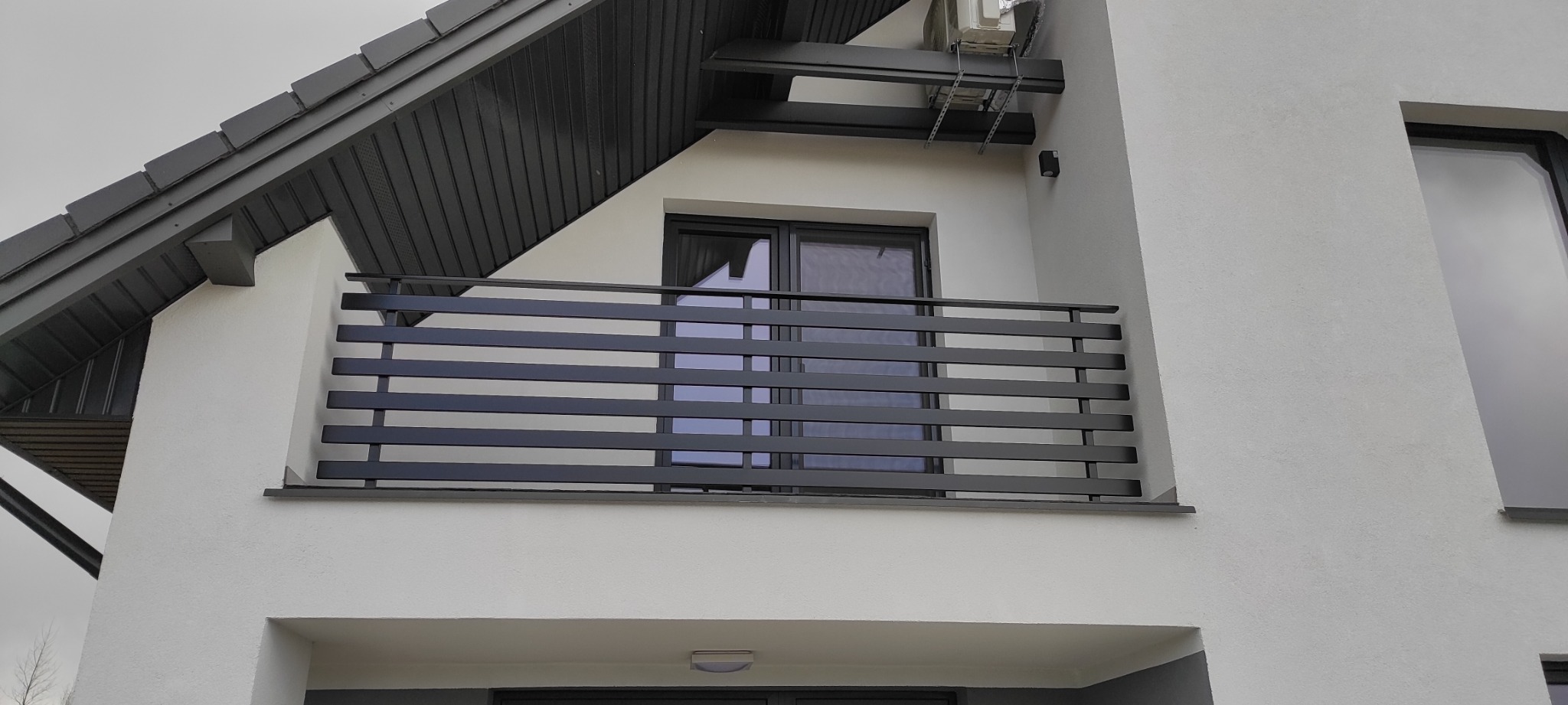 Nowoczesna, pozioma, czarna barierka balkonowa na tle białej elewacji domu z ciemną stolarką okienną i dachem; widoczny fragment klimatyzatora.