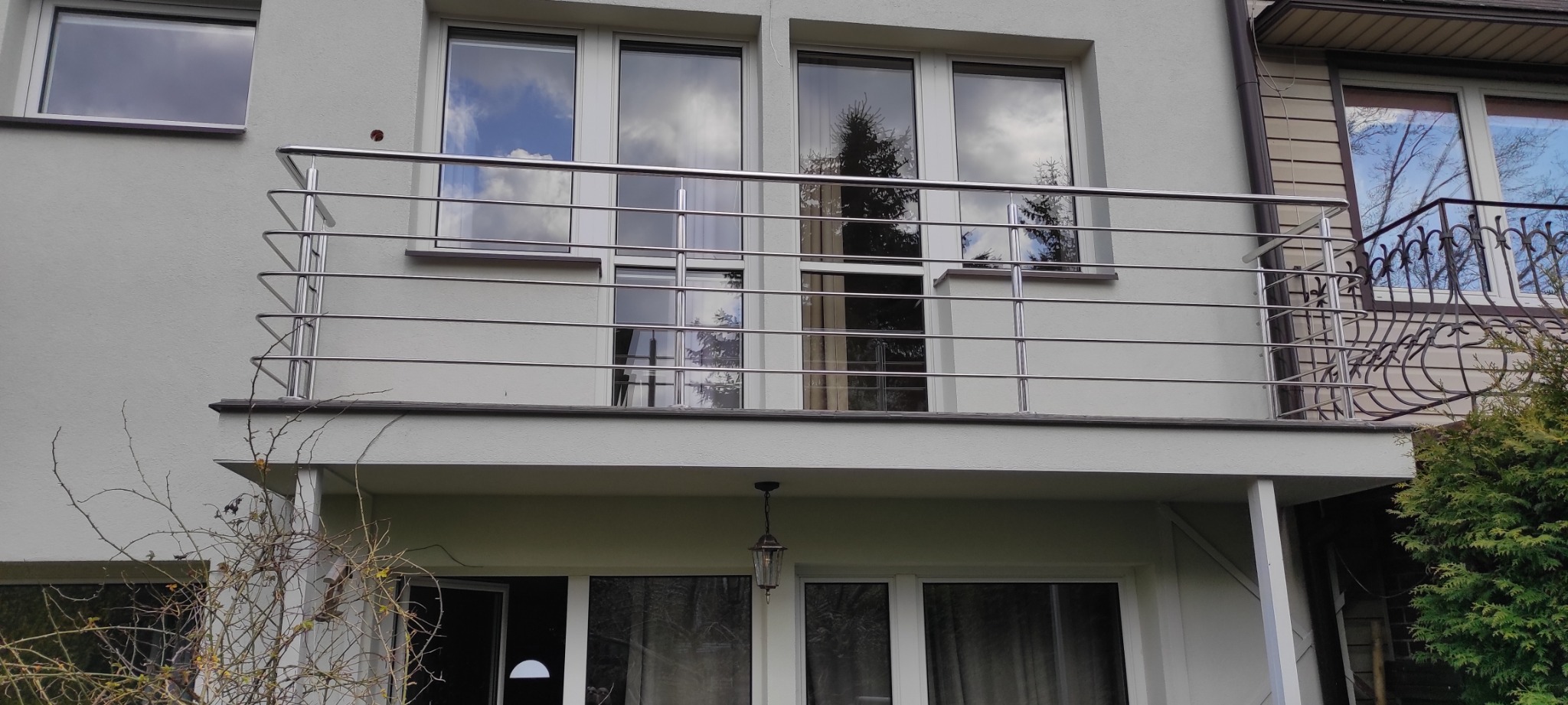 Balkon z nowoczesną, poziomą balustradą ze stali nierdzewnej, odbicia chmur i drzew w oknach, widok z dołu na elewację budynku.