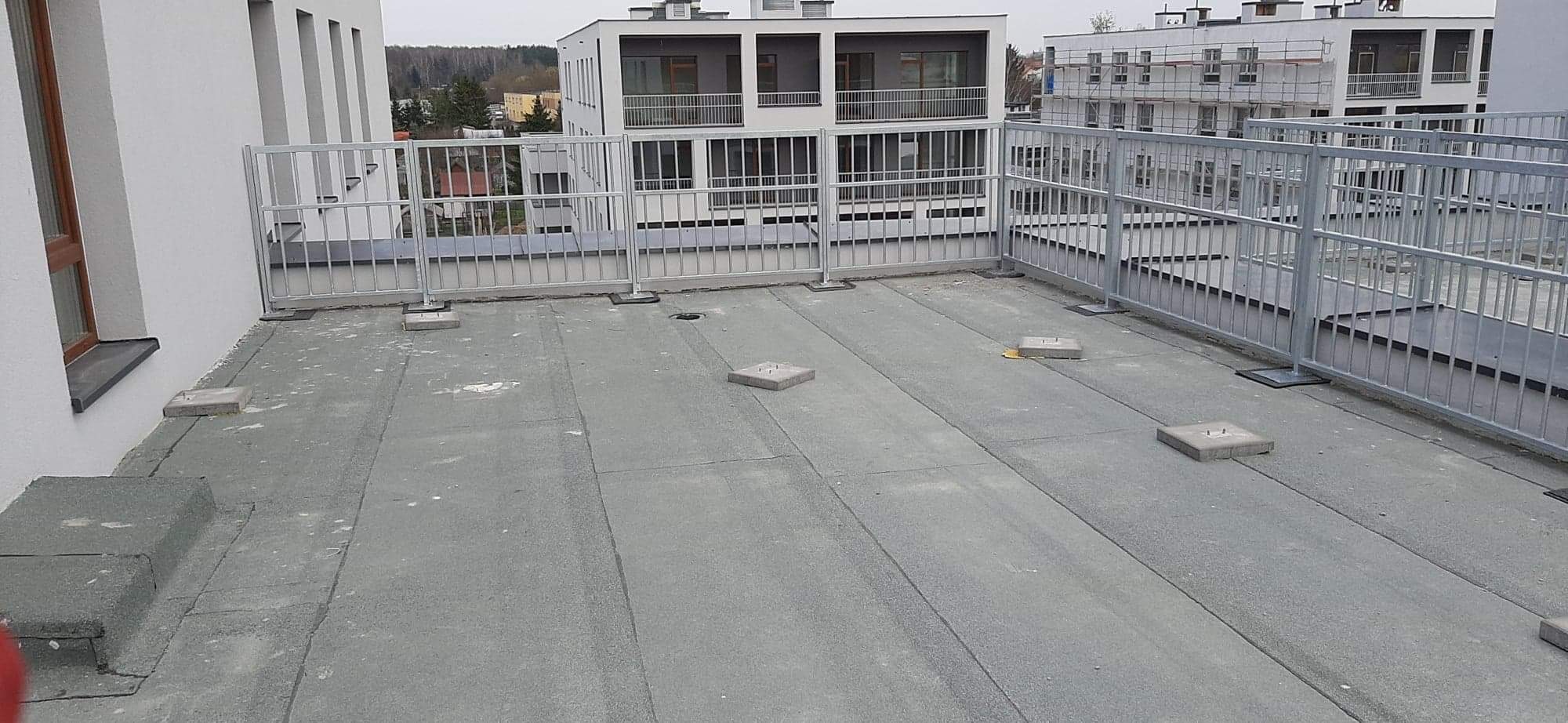 Szara, metalowa barierka ochronna zamontowana na płaskim dachu budynku wielorodzinnego, widoczne betonowe podstawy montażowe i fragmenty innych budynków w tle.