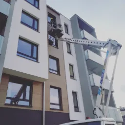 Pracownik w żółtym kasku na podnośniku koszowym podczas montażu lub naprawy okien w nowoczesnym budynku wielorodzinnym z brązową elewacją.