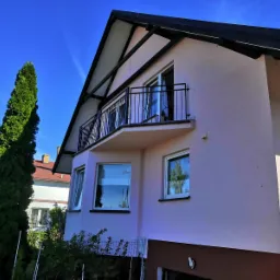 Jasnoróżowy dom z czarną, metalową balustradą balkonową w Kostrzynie nad Odrą, widziany z dołu pod kątem, z wysokim zielonym drzewem po lewej stronie.