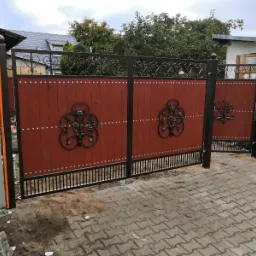 Dwuskrzydłowa brama wjazdowa z drewnianymi panelami w kolorze brązowo-czerwonym, zdobiona kutymi rozetami kwiatowymi i metalowymi nitami, osadzona na czarnej, metalowej ramie z poziomymi prętami u...
