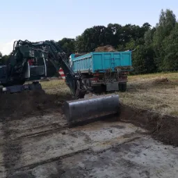 Ciemnozielona koparka Liebherr 314 z łyżką przed turkusową wywrotką wypełnioną ziemią na wyznaczonym terenie budowy w słoneczny dzień.