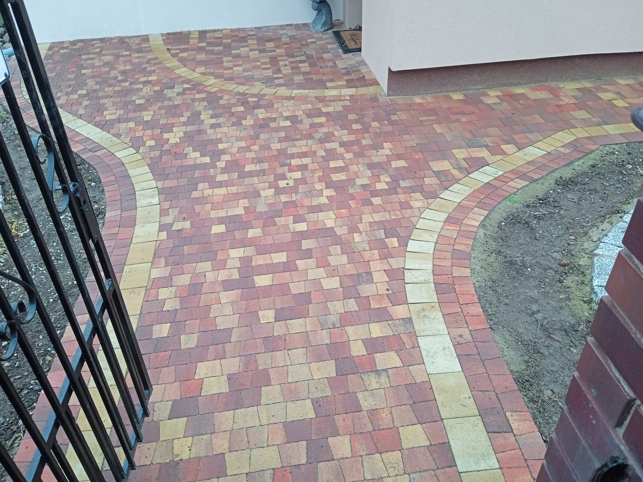 Ułożona kostka brukowa w odcieniach czerwieni i beżu, tworząca wzór na podjeździe. Widoczna brama z kutego żelaza i fragment elewacji budynku.