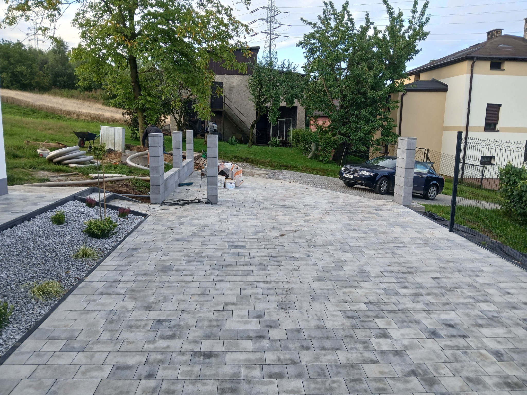 Szara kostka brukowa ułożona na podjeździe, z elementami dekoracyjnymi z kamienia i roślin. W tle budynek i samochód osobowy. Częściowo widoczne przygotowania do dalszych prac.