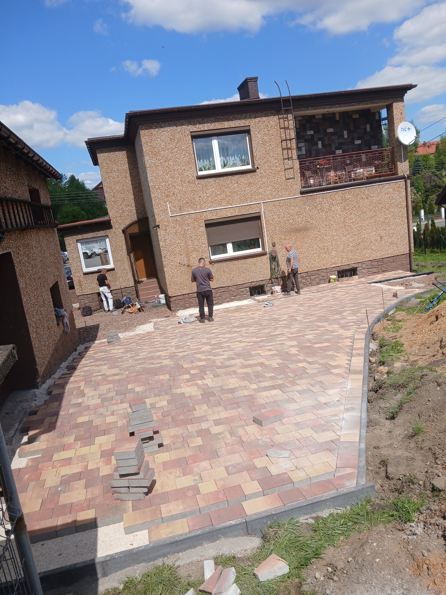 Układanie kostki brukowej w odcieniach czerwieni i beżu na podjeździe przed domem. Widoczni pracownicy i narzędzia. Częściowo ułożona kostka, krawężniki.