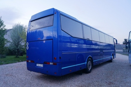 Tył niebieskiego autobusu turystycznego z widoczną tablicą rejestracyjną i elementami odblaskowymi, zaparkowanego na żwirowym podłożu, w tle budynek i drzewa.