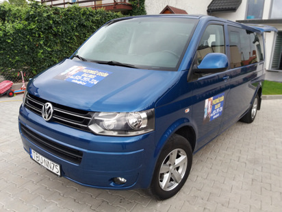 Niebieski Volkswagen Transporter z naklejką 'Przewóz Osób' na karoserii, zaparkowany przed budynkiem z zielonym żywopłotem w tle.