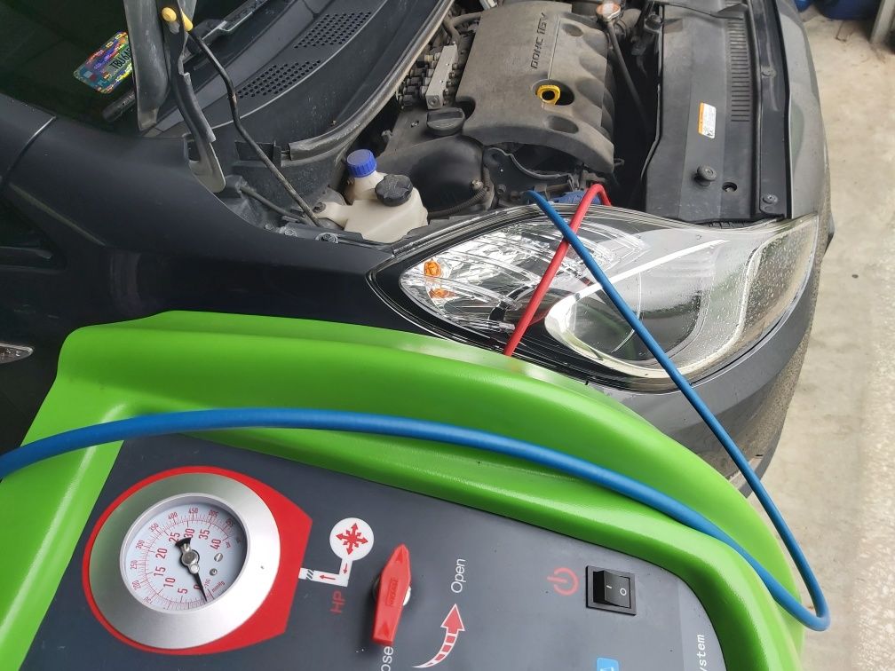 Podłączony sprzęt do klimatyzacji samochodowej przy otwartym silniku szarego auta, widoczne przewody czerwony i niebieski.