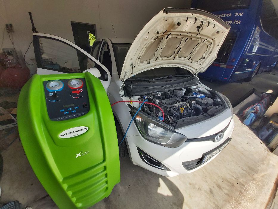 Biały samochód z otwartą maską, podłączony do zielonego urządzenia VIAKEN X520 do napełniania klimatyzacji, z otwartymi drzwiami kierowcy i widocznym autobusem w tle.