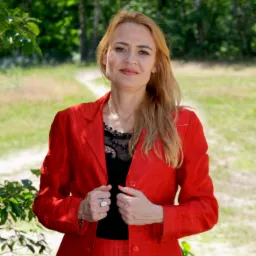 Kobieta w czerwonym kostiumie na tle zieleni, z widoczną ścieżką w oddali, uśmiechnięta, z rękami na klapach marynarki, prezentująca profesjonalny wizerunek.