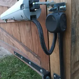 Automatyka bramy wjazdowej z napędem liniowym, widoczne elementy mocujące i przewody w peszlu, zamontowane na drewnianym skrzydle bramy.