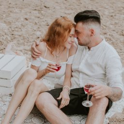 Ulotne chwile- Filipiak fotografia - Para siedząca na plaży, trzymająca kieliszki z czerwonym winem, w tle biała skrzynka i lampion.