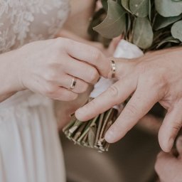 Ulotne chwile- Filipiak fotografia - Panna młoda zakłada obrączkę na palec pana młodego, trzymającego bukiet ślubny z białymi wstążkami.