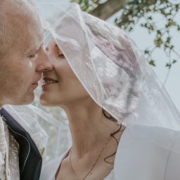 Ulotne chwile- Filipiak fotografia - Pocałunek nowożeńców w plenerze, panna młoda w welonie z subtelnym makijażem, widoczne fragmenty drzew i nieba w tle.