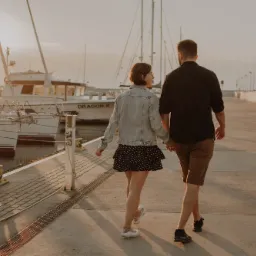 Para trzymająca się za ręce spaceruje po nabrzeżu portowym w ciepłym świetle zachodzącego słońca, w tle jachty i latarnie.