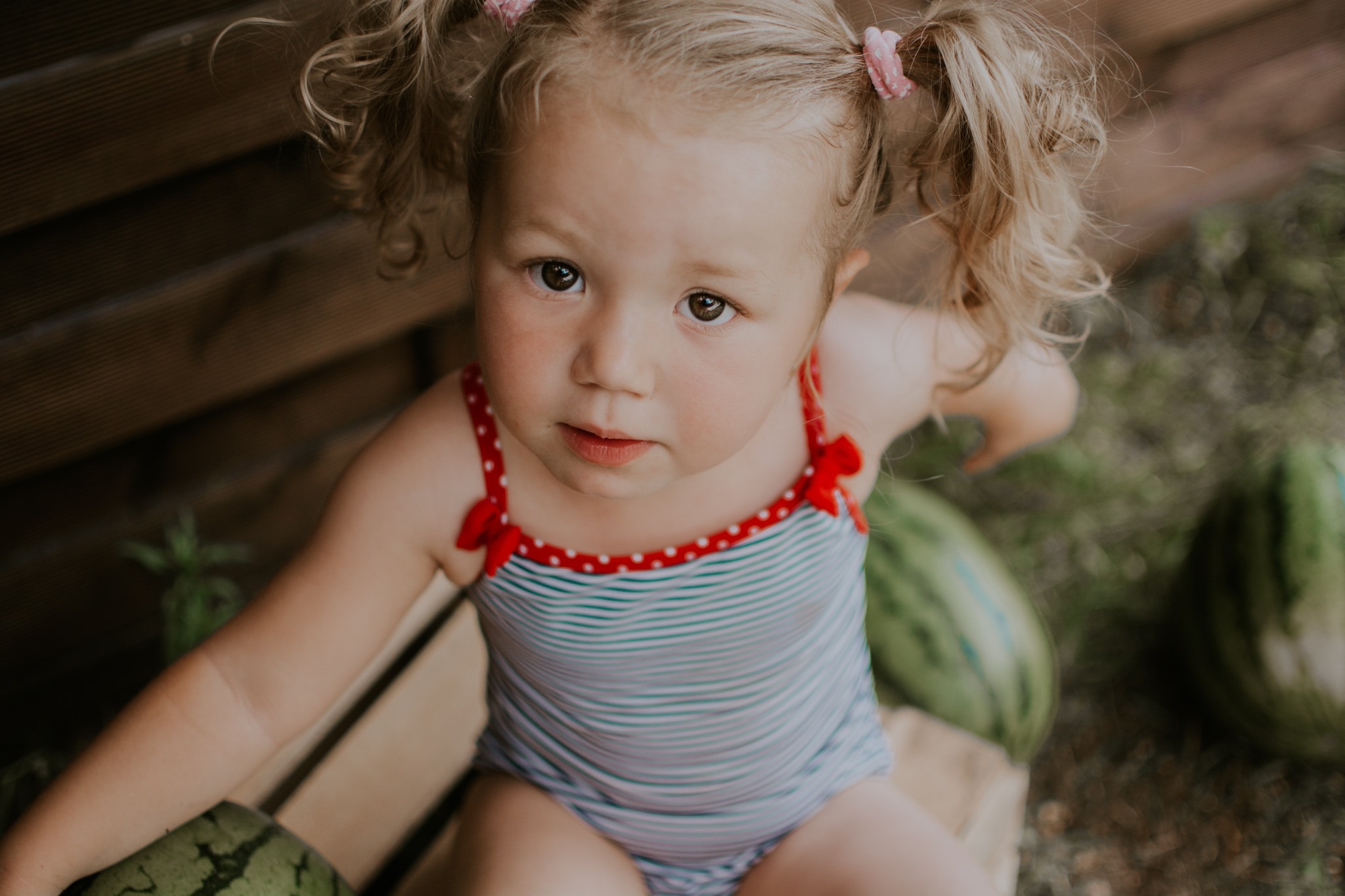 Dziewczynka w stroju kąpielowym w paski, siedząca na drewnianej ławce z arbuzami, z włosami spiętymi w dwa kucyki z różowymi gumkami, patrzy w górę.