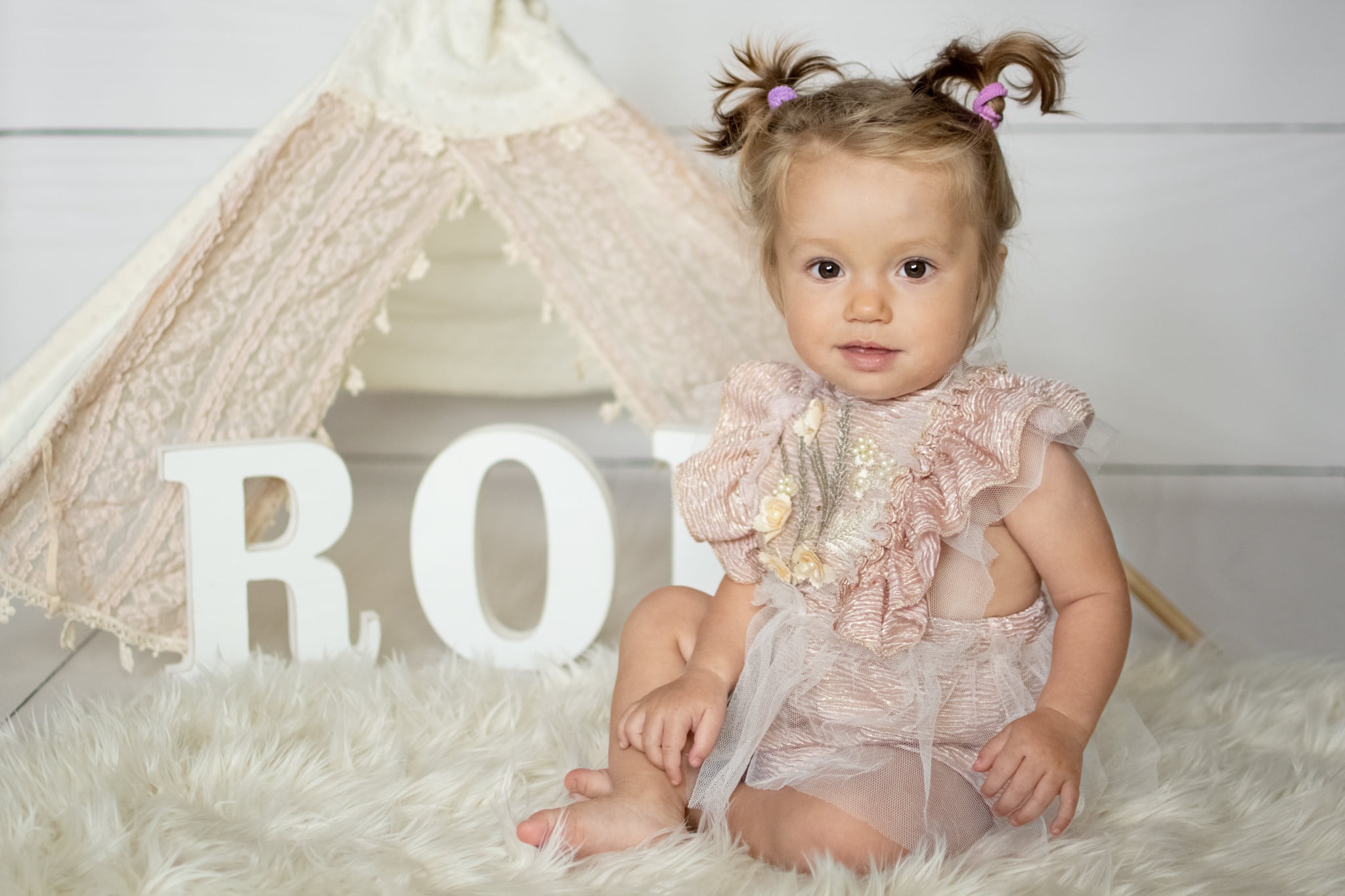 Mała dziewczynka w różowym stroju siedzi na białym futrze przed tipi i dużymi białymi literami tworzącymi słowo 'RO'
