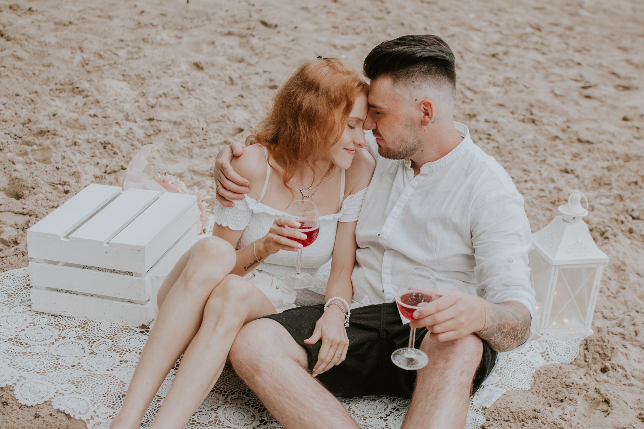 Para siedząca na plaży, trzymająca kieliszki z czerwonym winem, w tle biała skrzynka i lampion.