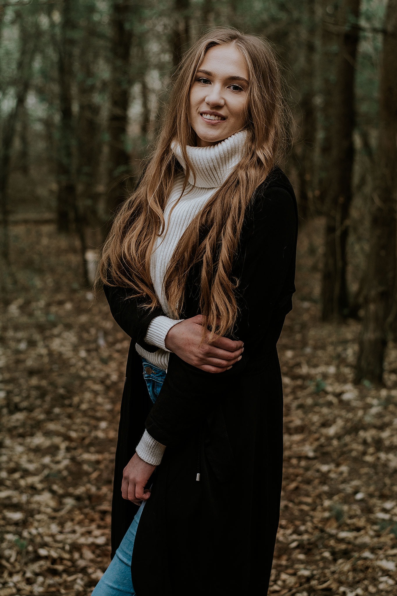 Portret uśmiechniętej kobiety w białym golfie i czarnym płaszczu na tle jesiennego lasu.