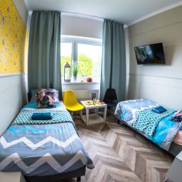 Pokoje Hotelowe Siedlczanka - Pokój dwuosobowy w jasnych barwach z dwoma łóżkami, żółtym akcentem krzesła i stolikiem z poczęstunkiem, wzorzysta tapeta na ścianie i telewizor LCD.